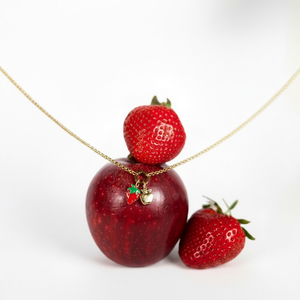 Nadare Co. Apple Charm and strawberry charm on a gold chain draped over a fresh red apple and strawberries.